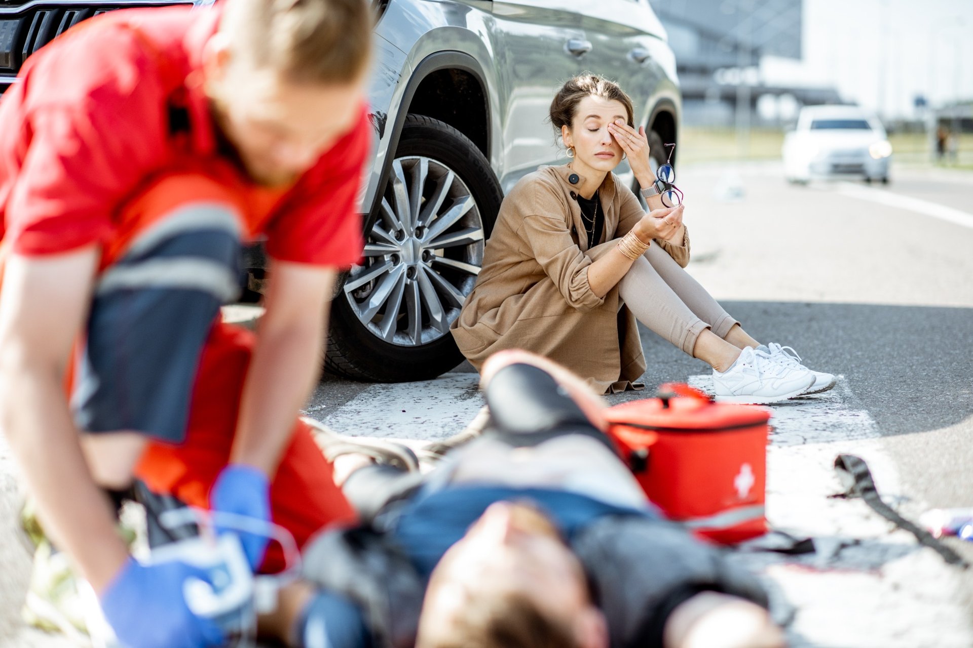 Distressed woman sitting by her damaged vehicle while a medic assists an injured person on the road