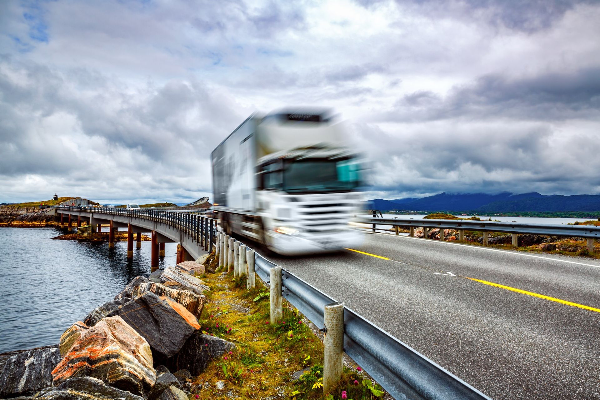 Motion-blurred semi-truck speeding across a scenic coastal bridge under dramatic skies