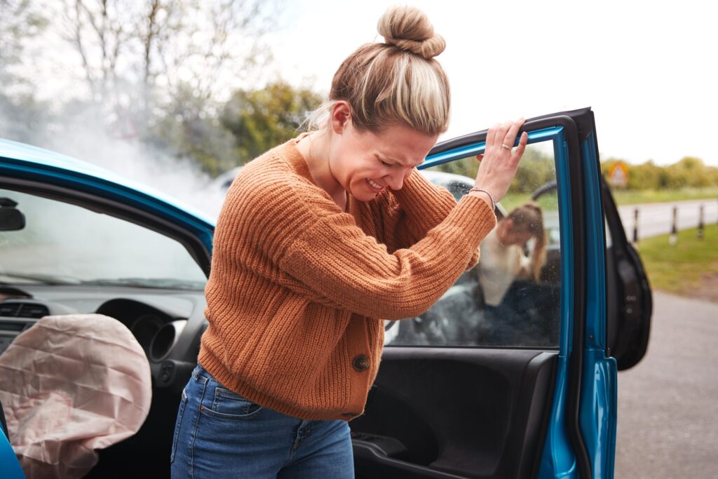 Female driver in an orange sweater clutching her head in pain while exiting a blue car with a deployed airbag after a road accident